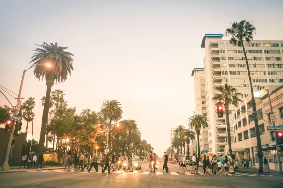 Busy pedestrian crosswalk on wide boulevard in Santa Monica, California lined with tall palm trees against golden hour light.